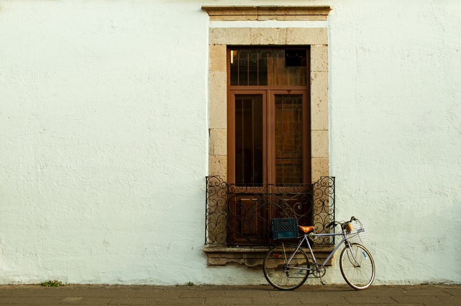 A classic bicycle rests against a textured wall beside a traditional Mexican window on a quiet street.