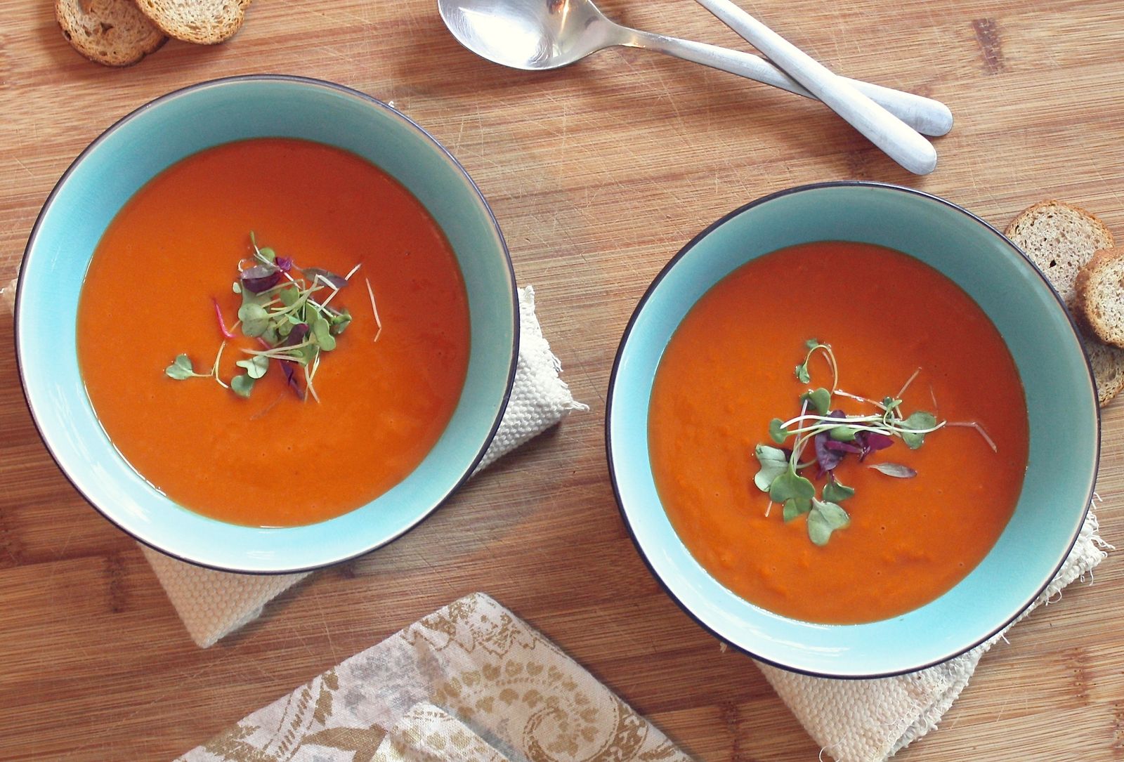 Two teal bowls of tomato soup topped with microgreens on a wooden table
