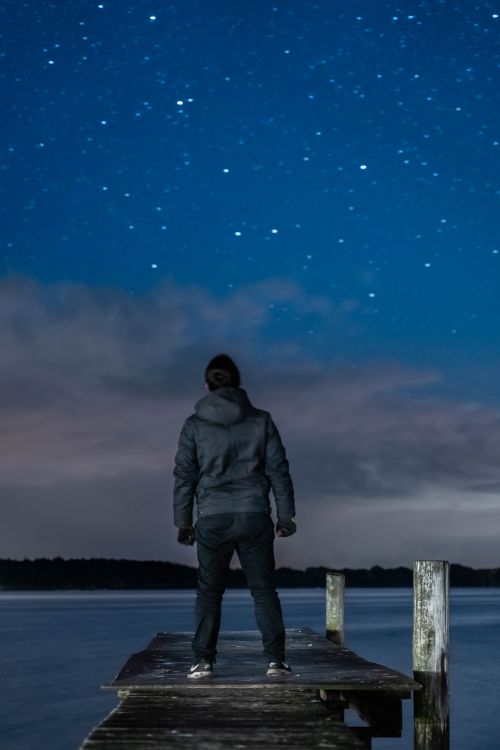 Free A person stands on a wooden dock, gazing at a starry night sky over a serene lake. Stock Photo