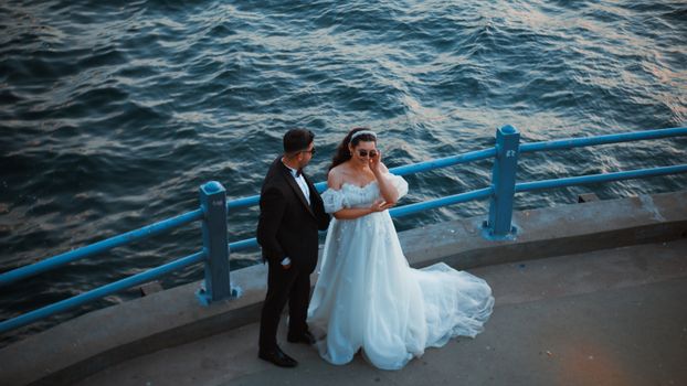 Bride and groom in wedding attire standing romantically by the seaside promenade.