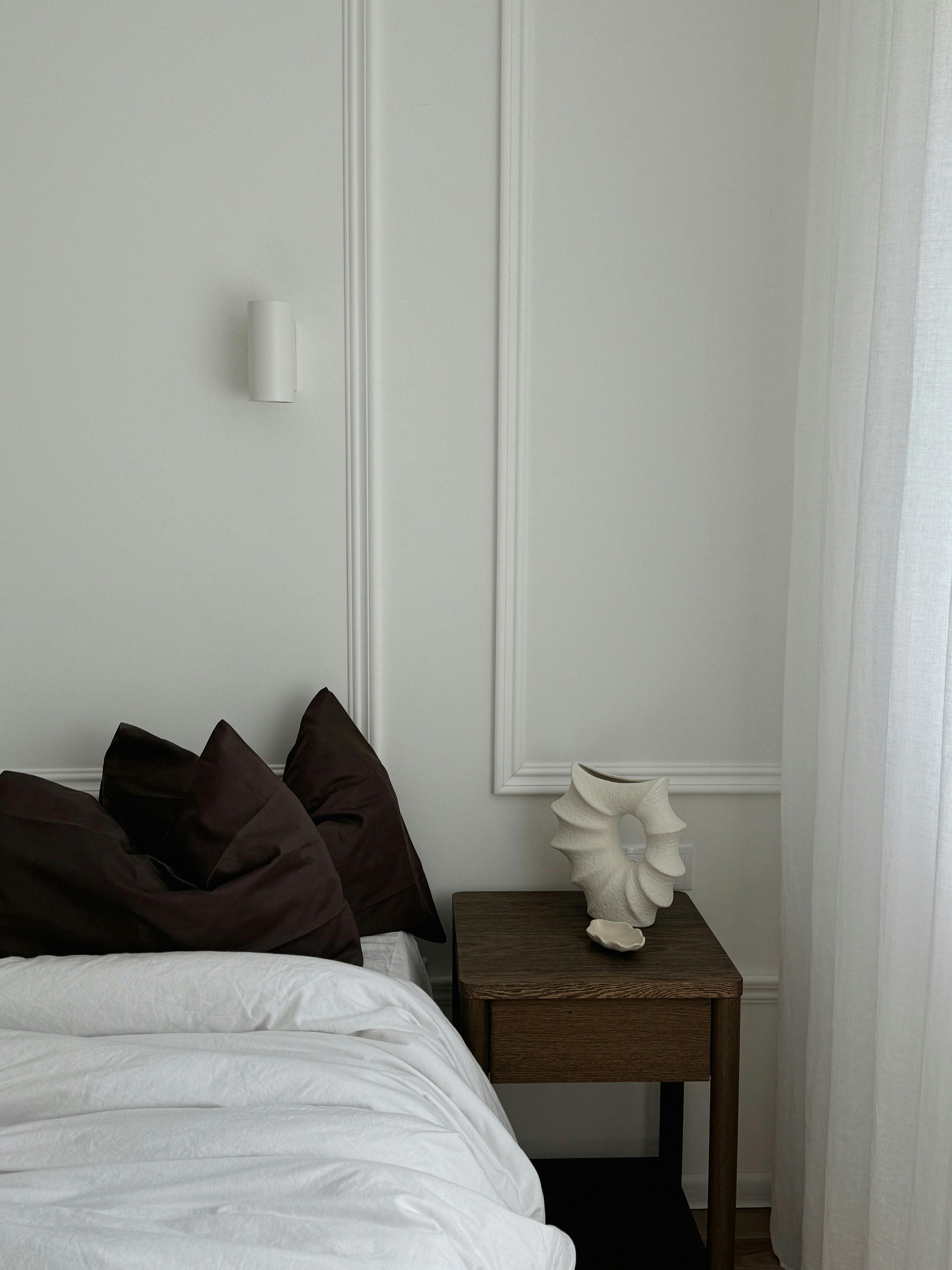 A minimalist bedroom corner with dark pillows, nightstand, and ceramic sculpture.