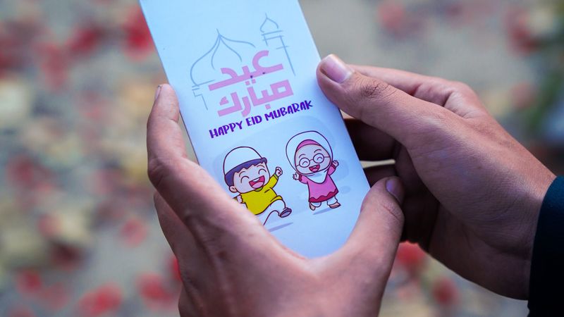 Close-up of a person holding an Eid Mubarak card with festive design outdoors.
