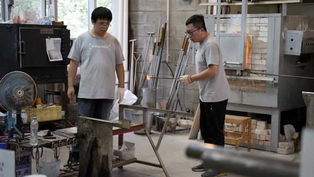 Two artisans intently engaged in glassblowing inside a lively workshop environment. - Fotografer Sidoarjo
