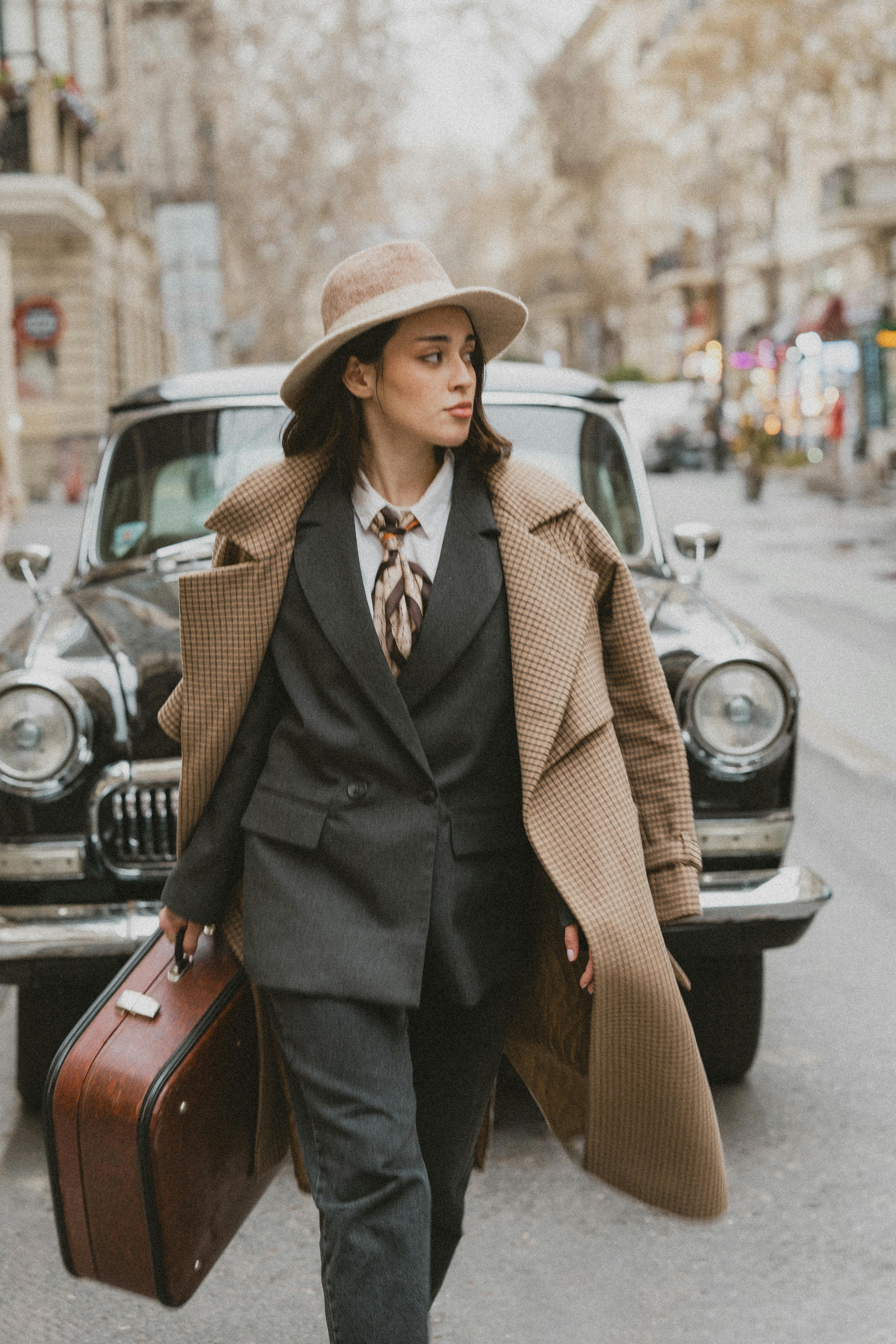 a woman in a suit and hat walking down the street with a suitcase