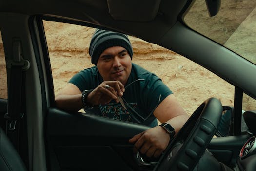 Casual man wearing a beanie leans on car window holding sunglasses outdoors.