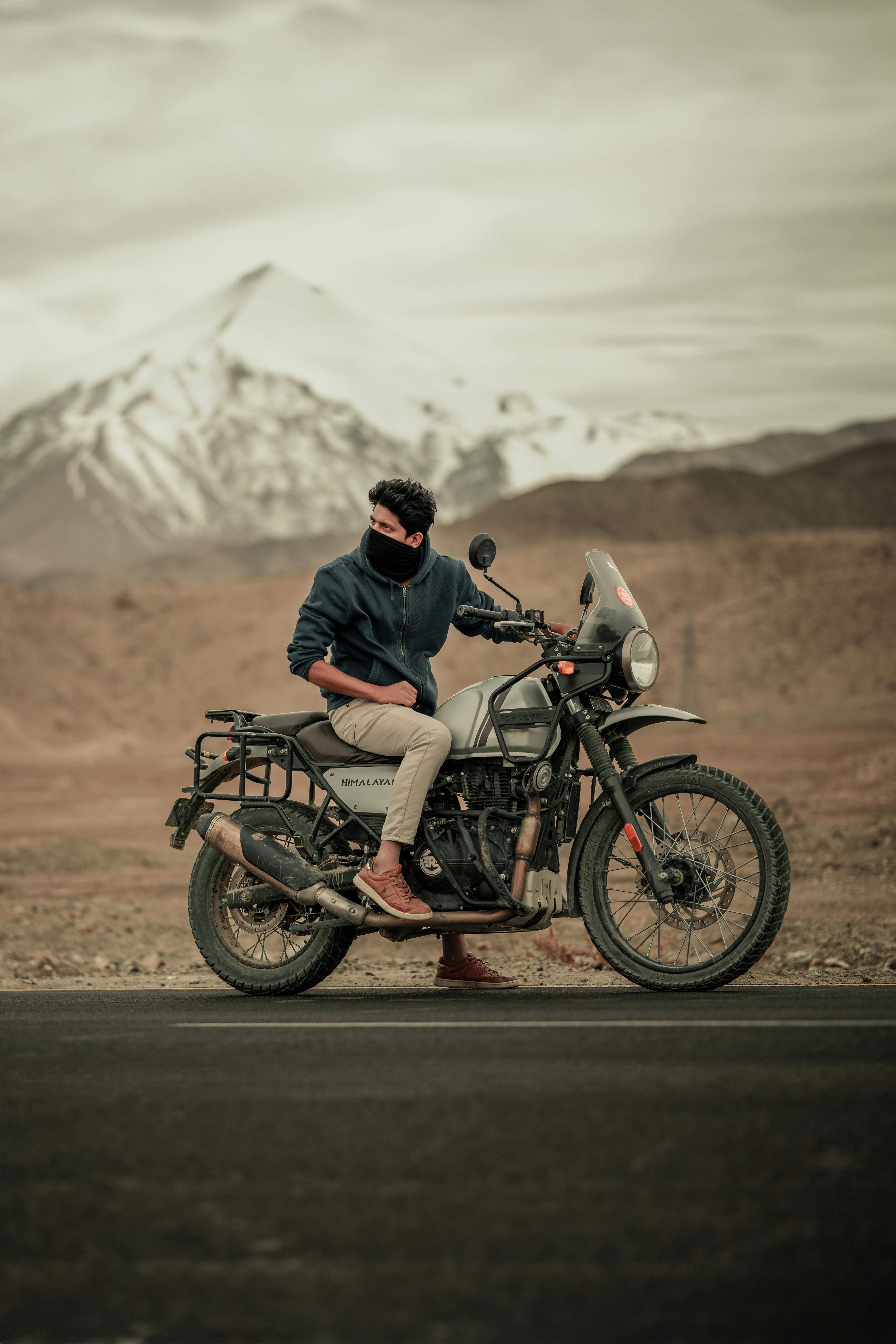 A biker with a mask sitting on a motorbike near the mountains in India, showcasing adventure travel.