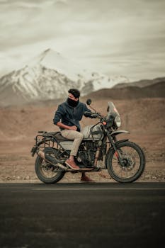 A biker with a mask sitting on a motorbike near the mountains in India, showcasing adventure travel.