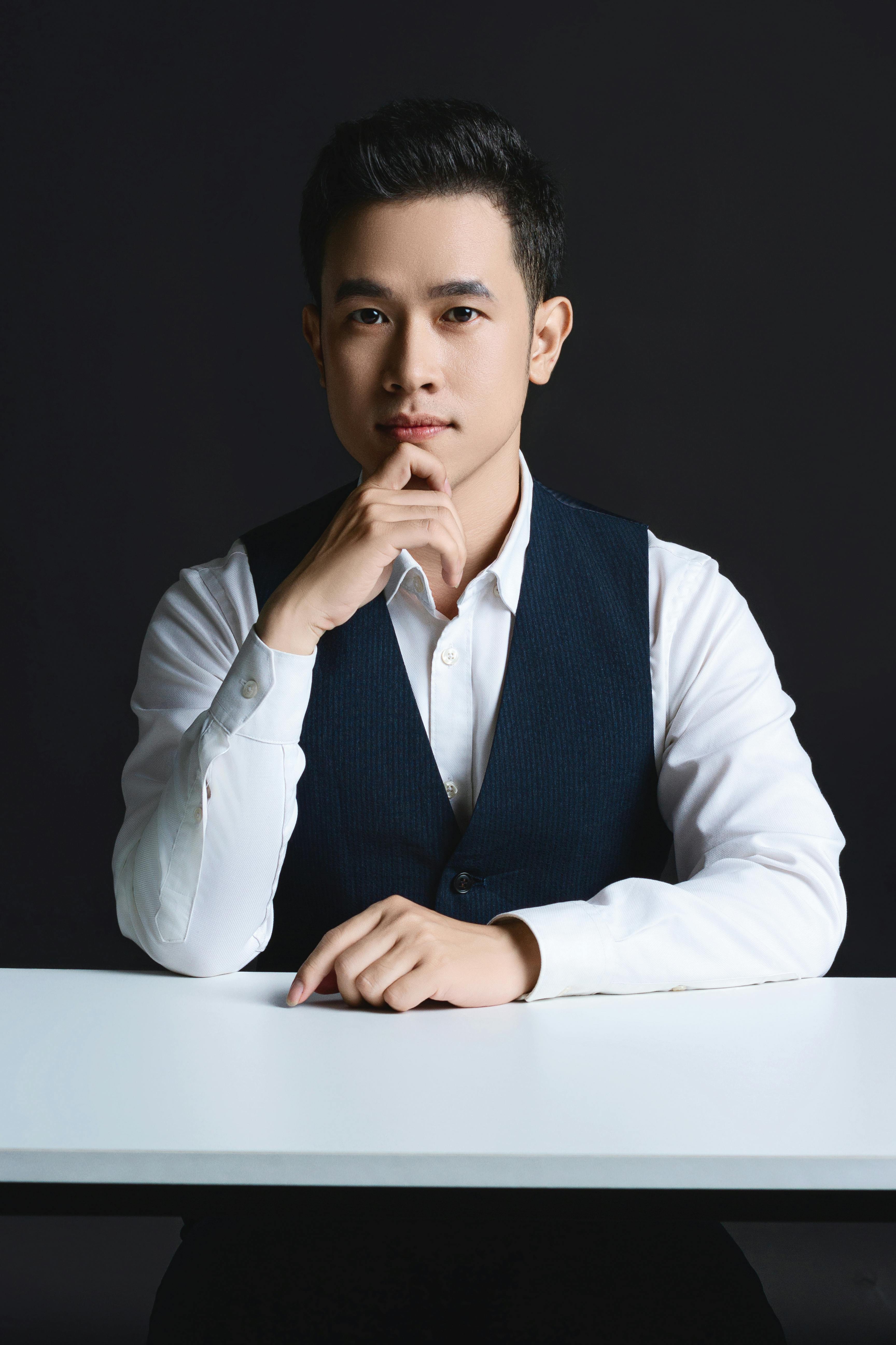 Elegant studio portrait of a young, well-dressed Asian man in a suit sitting thoughtfully at a table.