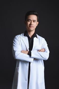Portrait of a confident Asian doctor in a white coat with arms crossed, set against a black background.