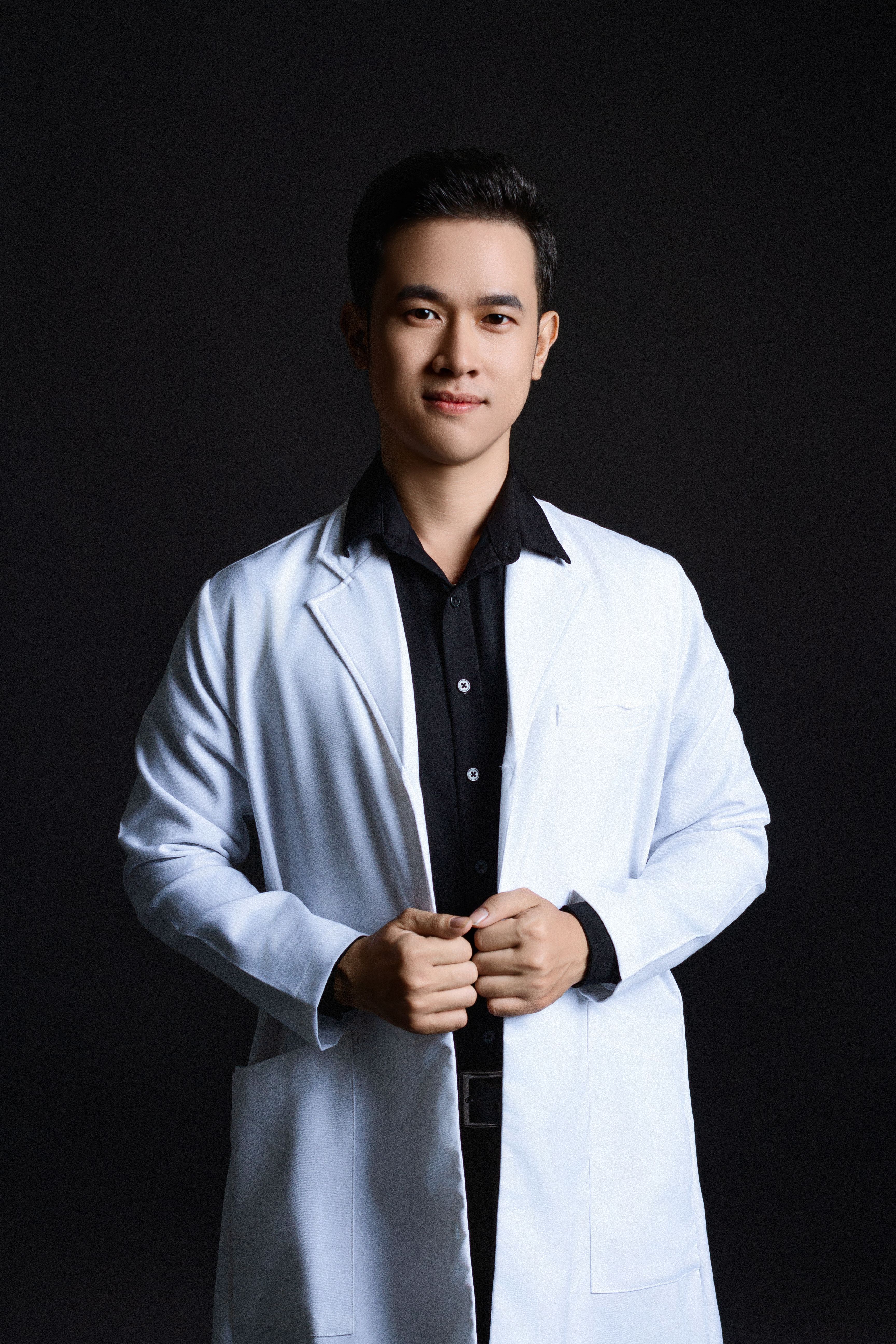 Portrait of a young male doctor in a studio setting, wearing a lab coat over a black shirt, exuding professionalism.