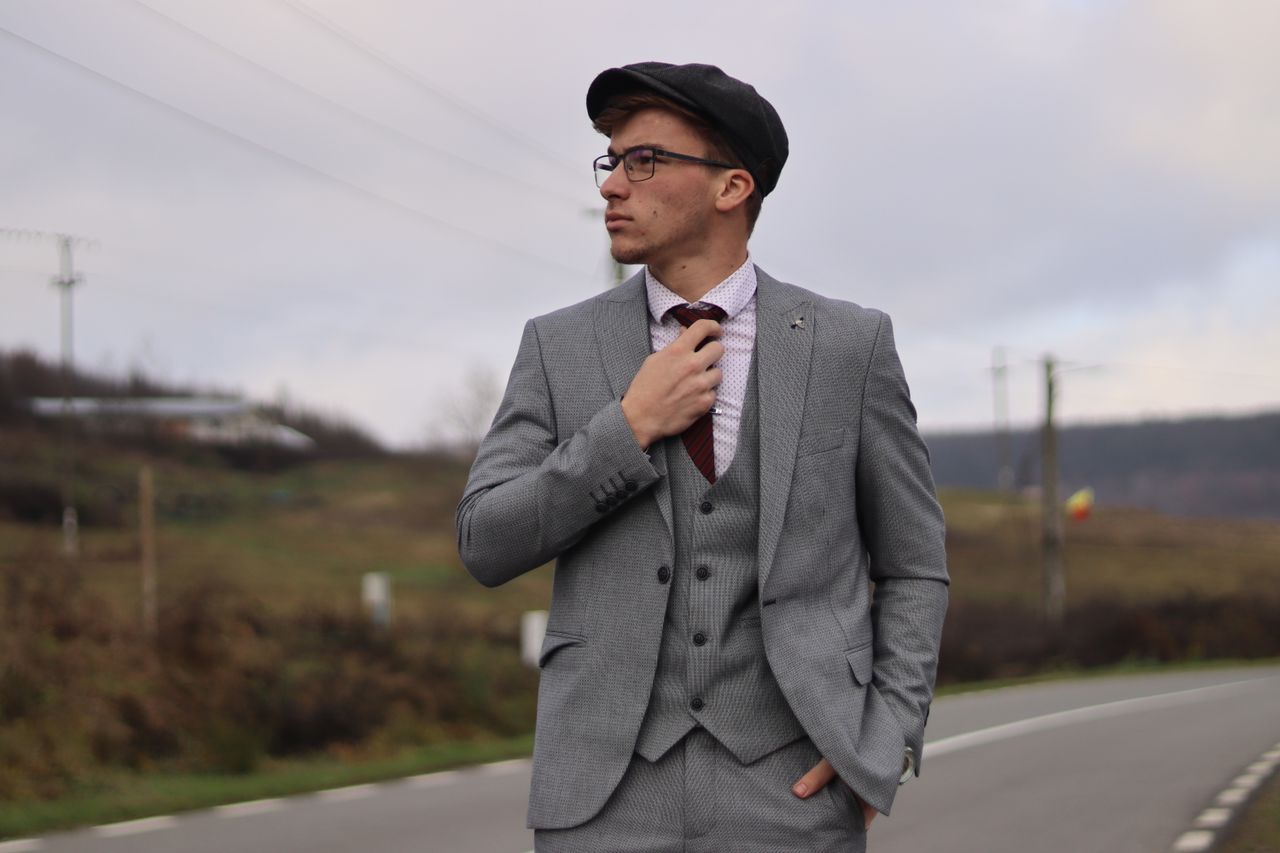 a fashionable man in a grey suit confidently poses on a rural road exu