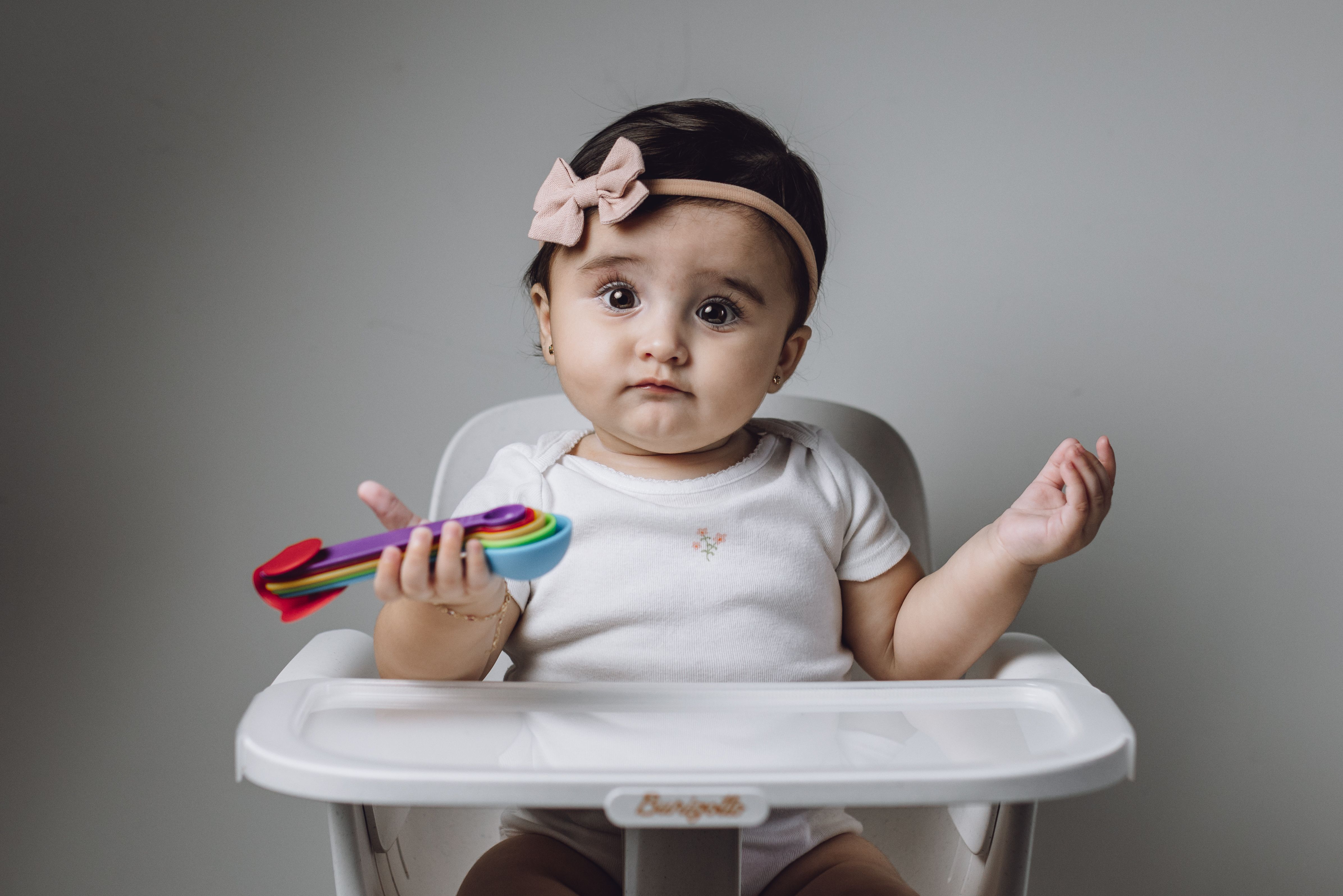 Cute baby girl in a high chair holding a colorful spoon, in a simple indoor setting.