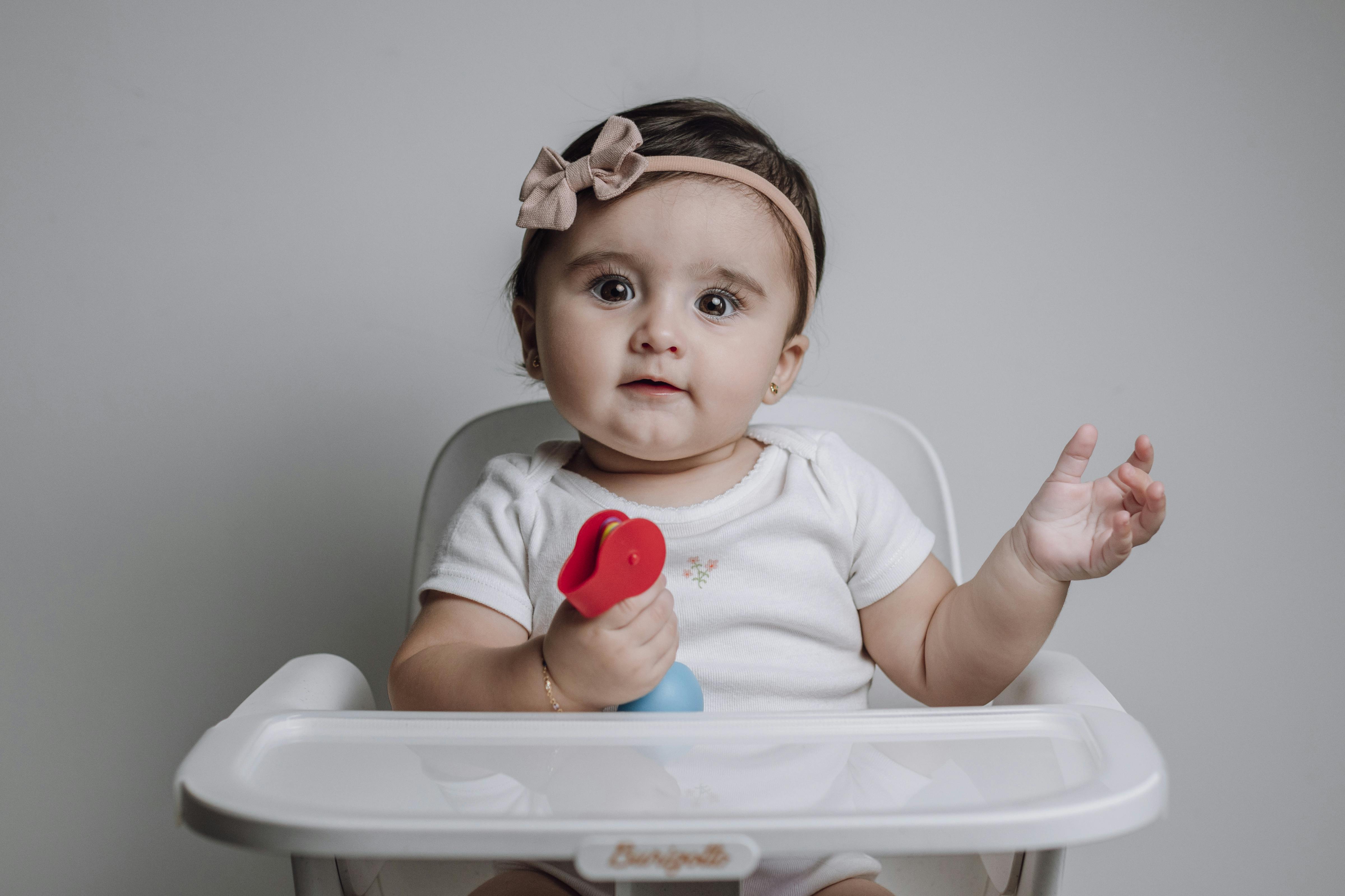 Cute baby girl with bow headband sitting in a high chair, playing with a toy.