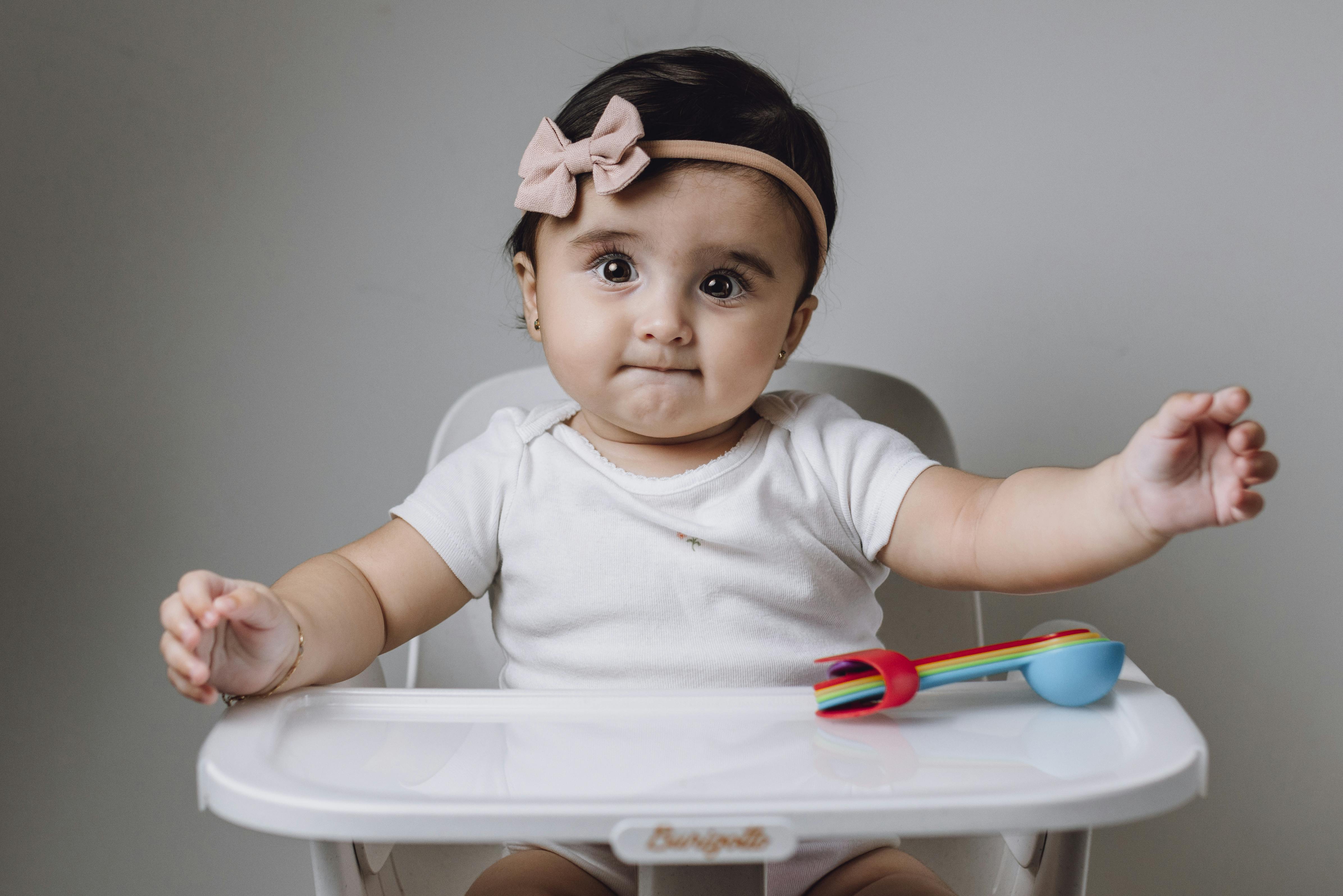 Cute baby girl in high chair with colorful toy utensils against gray background.