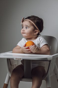 Charming portrait of a baby girl in a high chair, holding an orange with a thoughtful expression.