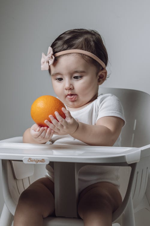 A baby girl sitting in a high chair holding an orange