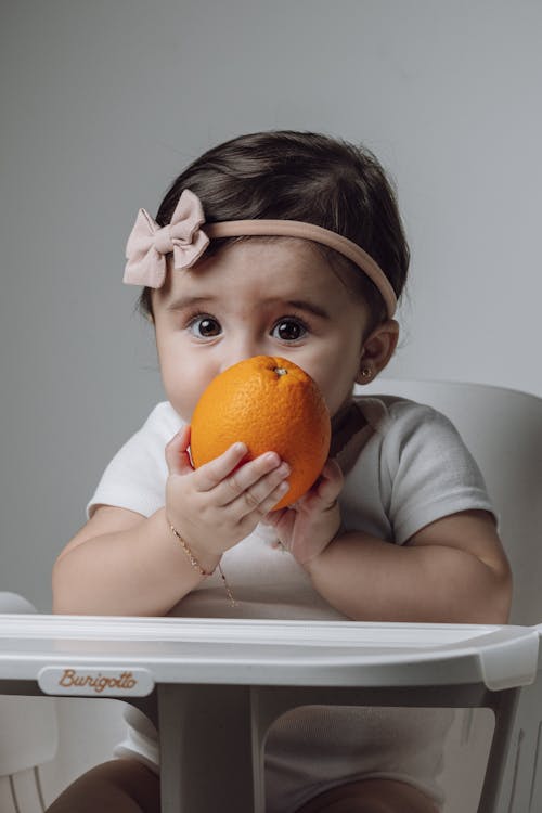 A baby girl is holding an orange in her high chair