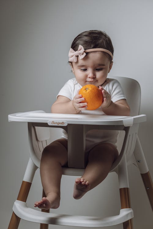A baby girl sitting in a high chair eating an orange