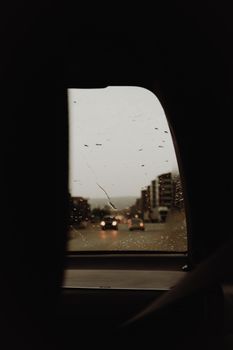 A rainy city street viewed through a car window with blurred cars and buildings.