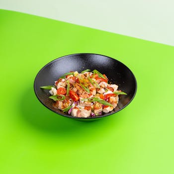A vibrant chicken salad with fresh vegetables in a black bowl against a green background.