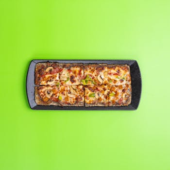Top view of square pizza slices with cheese and mushrooms on black tray against green background.
