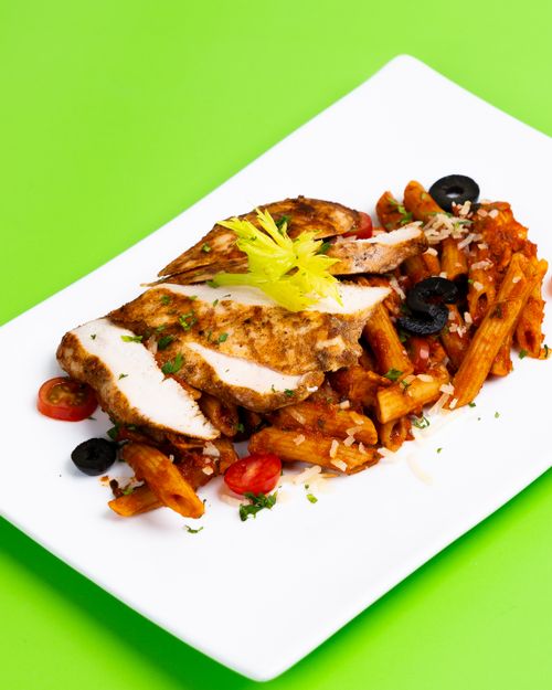 Free Delicious grilled chicken breast served with penne pasta, black olives, and cherry tomatoes on a vibrant green background. Stock Photo