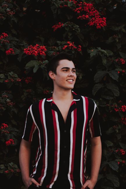 Young man in a striped shirt posing confidently with a smile amidst vibrant red flowers and lush greenery.