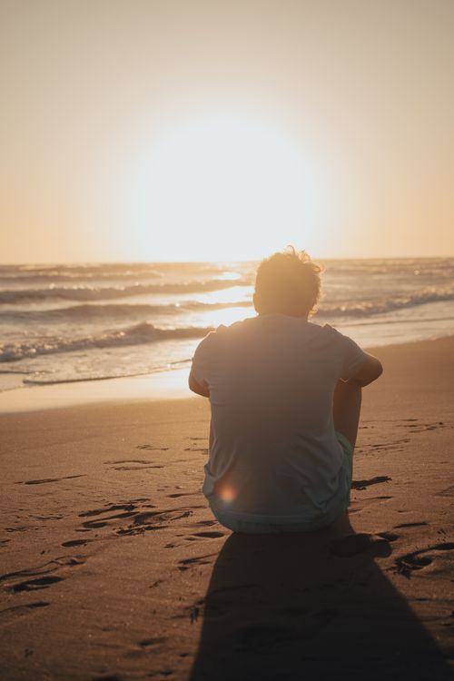Free Silhouette of a person reflecting while seated on a peaceful beach during sunset. Stock Photo
