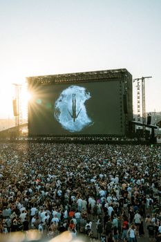 A vibrant outdoor music festival with a large crowd enjoying a performance against a beautiful sunset.