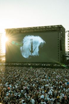 Massive crowd at outdoor music concert with a huge projection screen and sunlight flair.