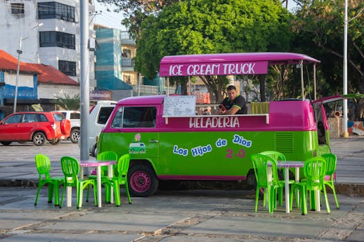 Bright ice cream truck serving customers on a sunlit urban street with colorful chairs.