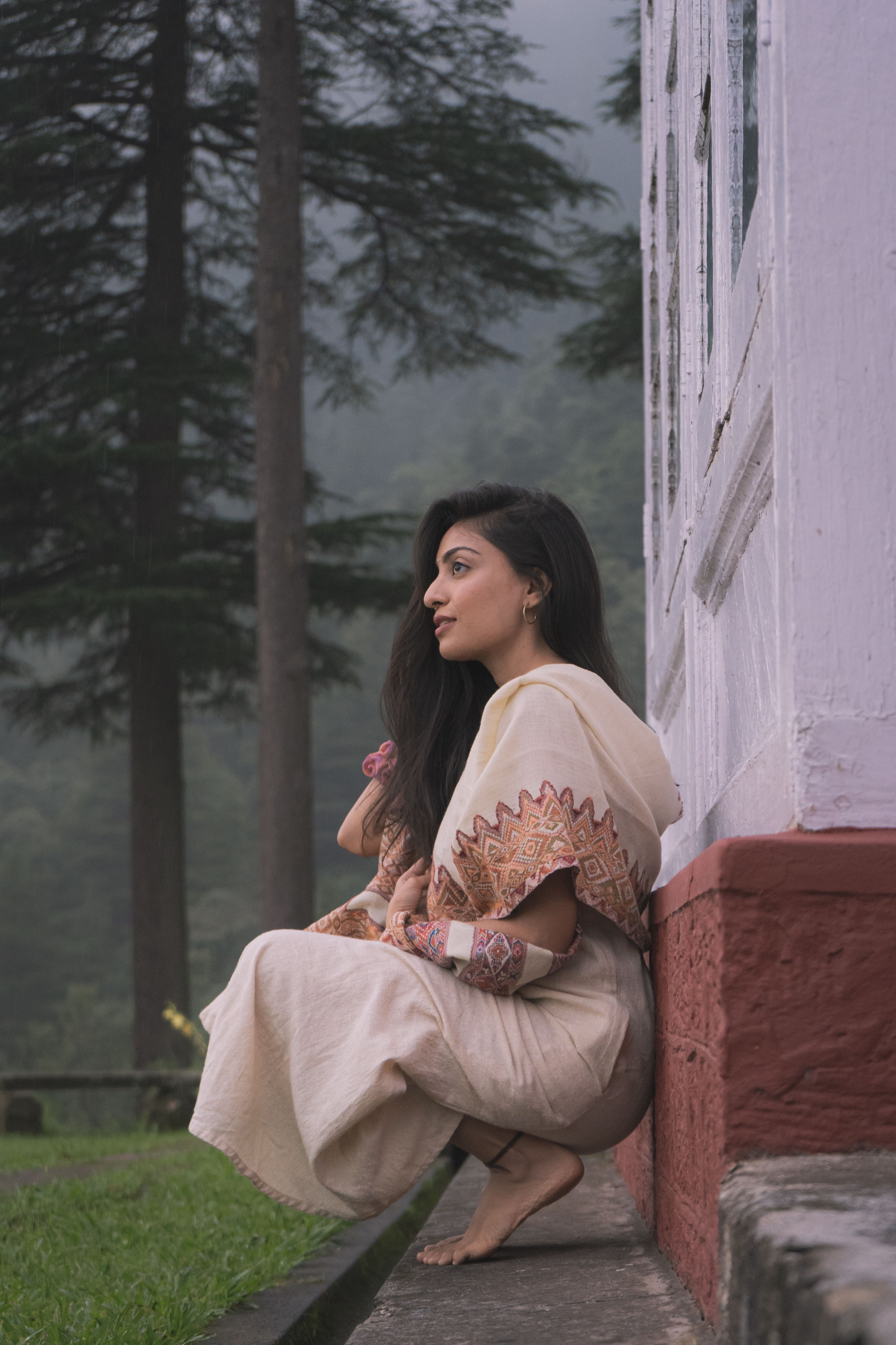 Woman crouching against a wall in a foggy garden in Dharamshala, India, surrounded by trees.