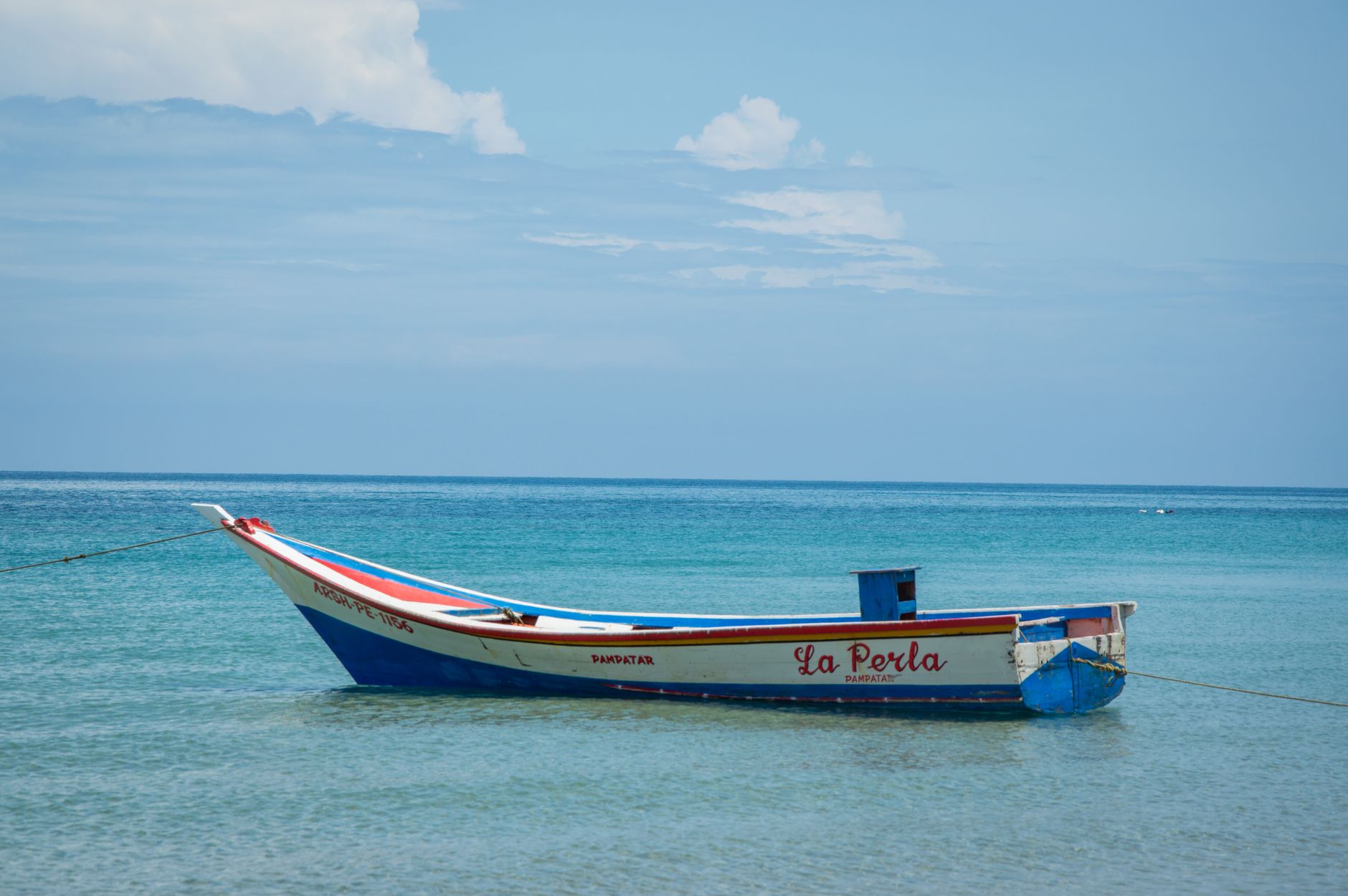 A traditional boat named La Perla moored on a tranquil, clear blue sea under a partly cloudy sky.