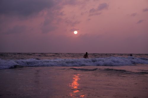 Free Silhouette of a person wading in the ocean at sunset with waves reflecting the sunset hues. Stock Photo