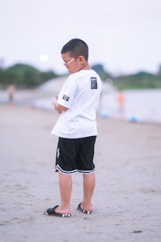 A young boy in casual wear stands on a serene beach, enjoying a quiet moment.