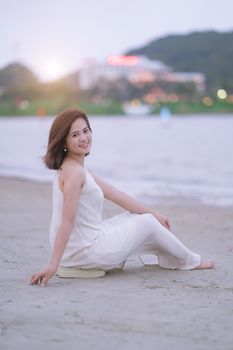 Woman in a white dress sitting on the beach, smiling at sunset.