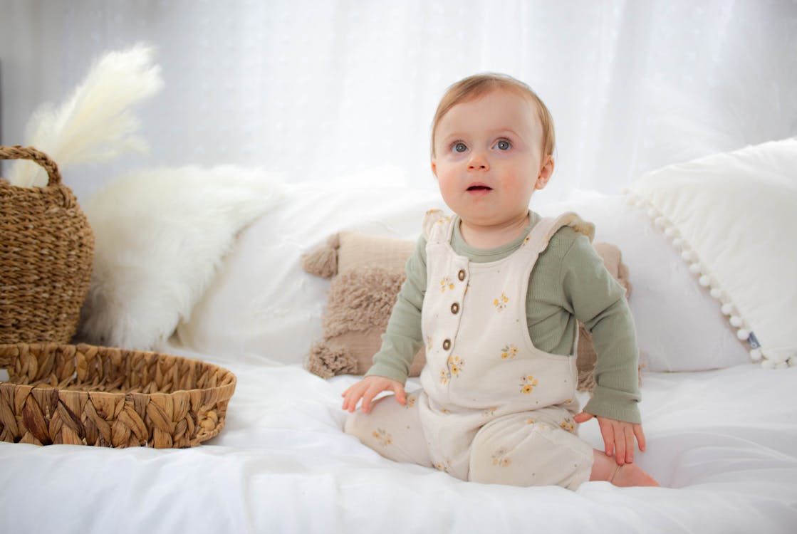 Free A baby sitting on a bed wearing a white overall Stock Photo