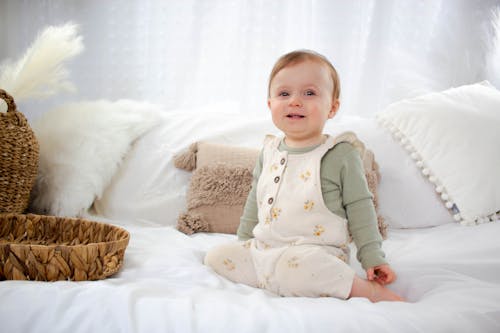 A baby sitting on a bed wearing a white overall
