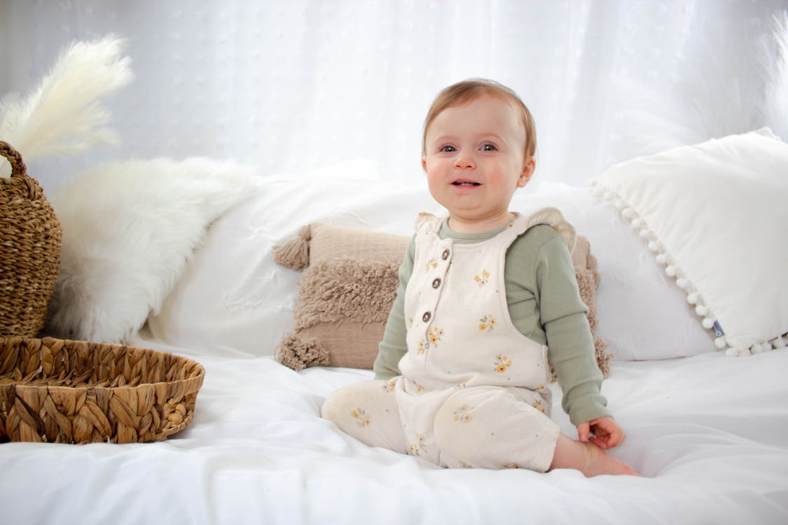 Free A baby sitting on a bed wearing a white overall Stock Photo