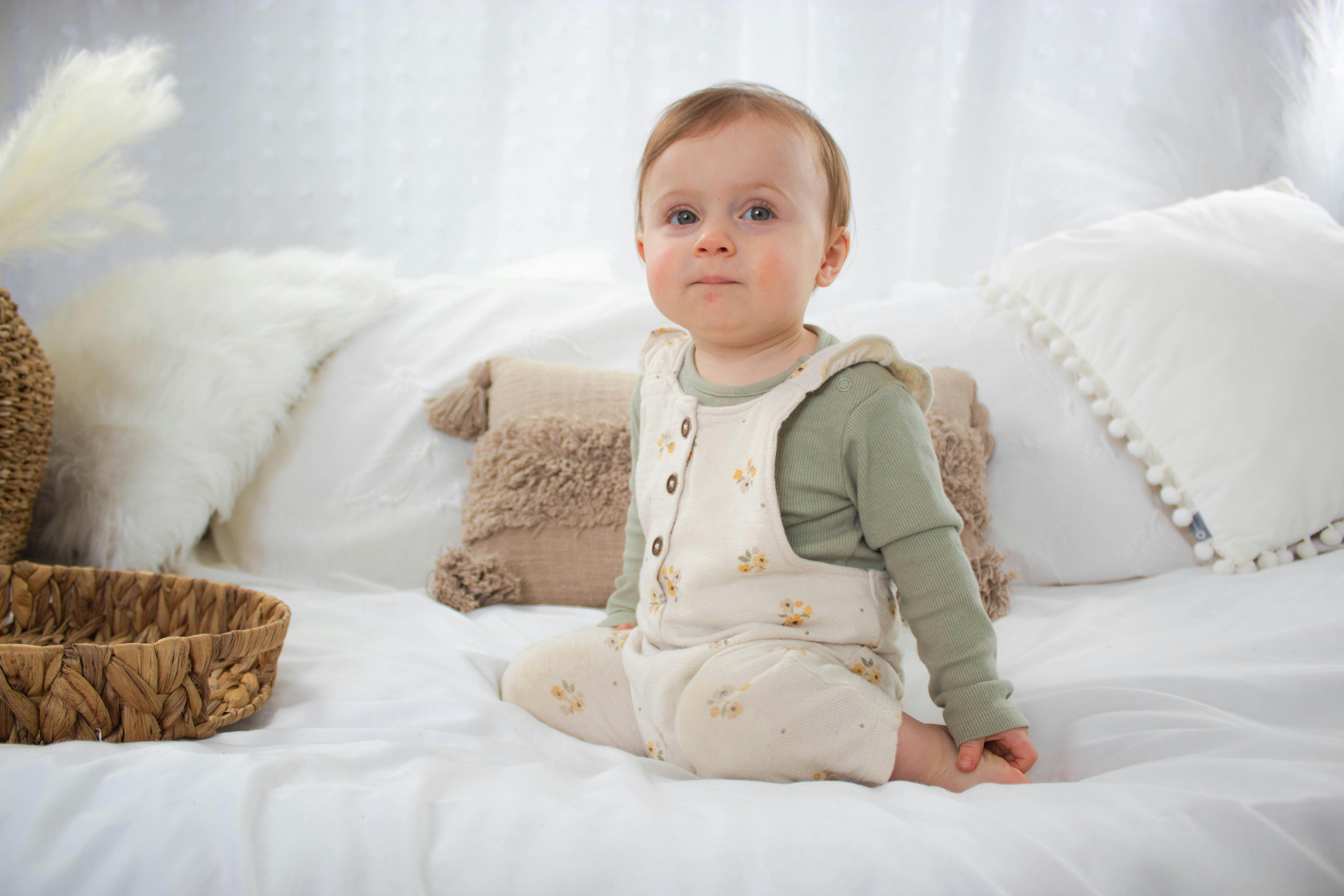 Adorable baby in soft pajamas sitting on a comfortable bed with pillows.