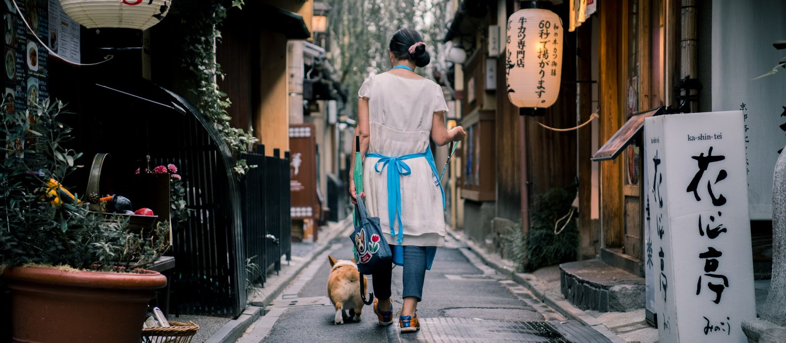 An Asian woman walks her dog through a traditional Kyoto alley, surrounded by lanterns and wooden houses.