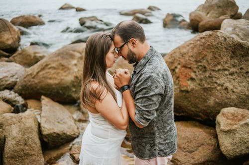Photo of Couple Holding Each Other While Standing by Rocks Next to Body of Water