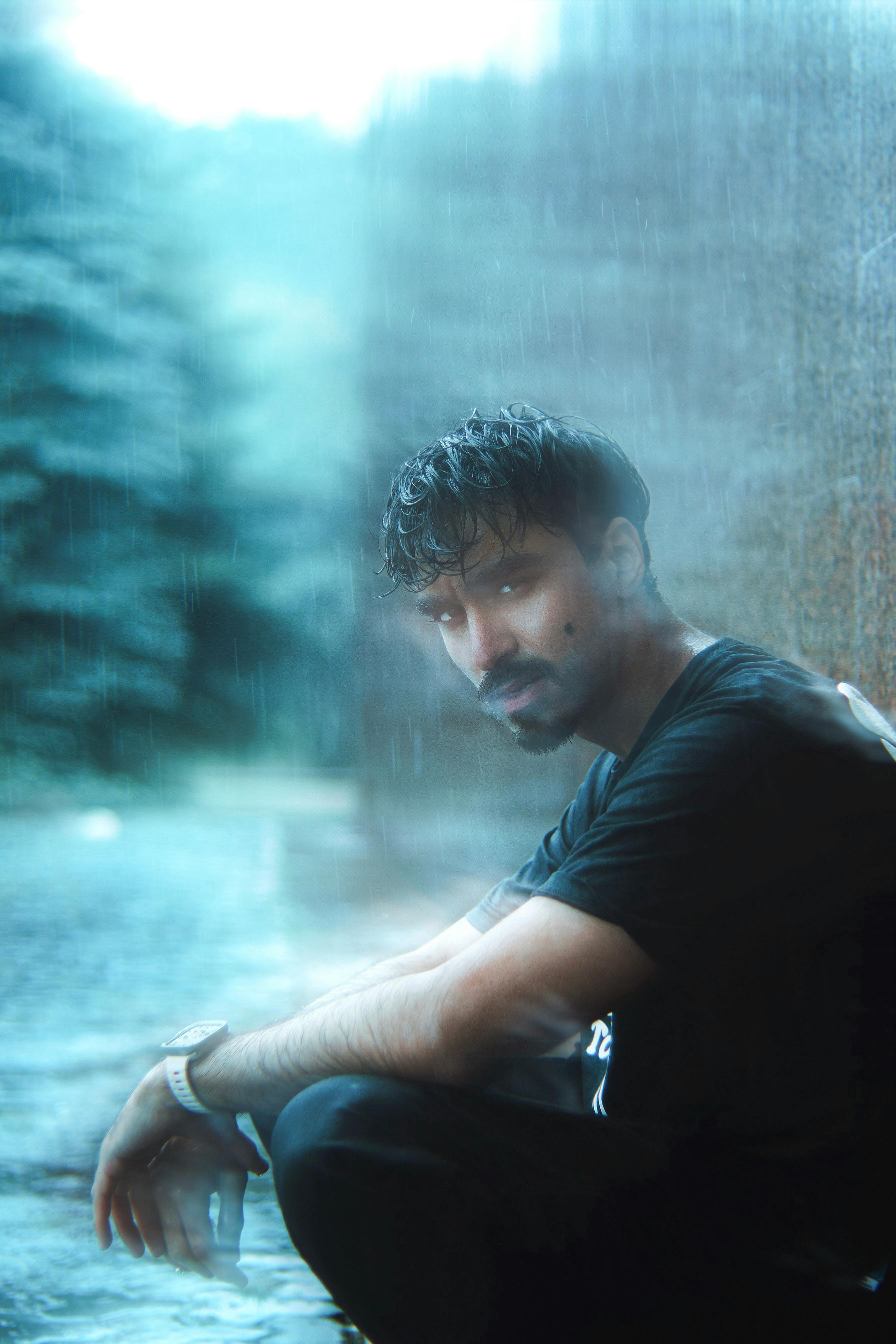 A young man with wet hair sits outdoors in the rain, lost in thought and introspection.