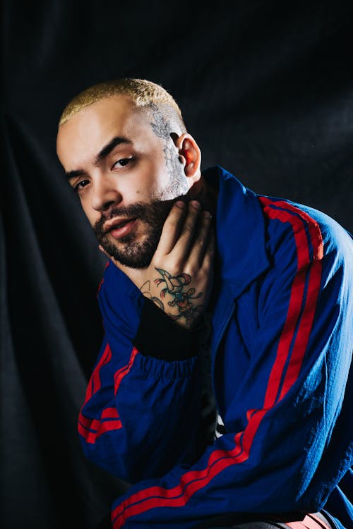 Photo of Tattooed Man in Blue and Red Jacket Posing In Front of  Black Background