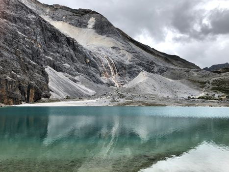 A tranquil turquoise lake reflecting rugged mountain slopes under a cloudy sky.