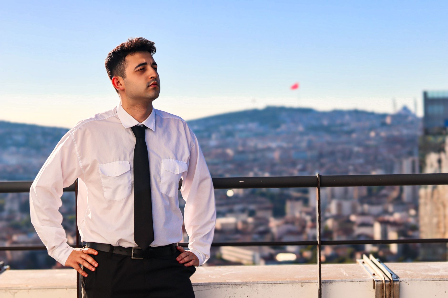 Portrait of a confident businessman standing with hands on hips on a rooftop terrace with a cityscape background.