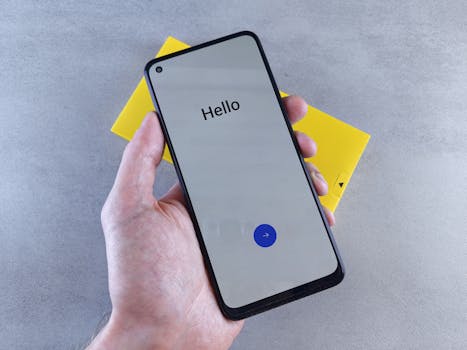 A hand holding a smartphone displaying 'Hello' on a gray surface with a yellow box underneath.