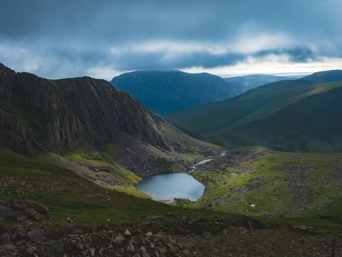 Free A tranquil lake nestled in a lush green valley surrounded by dramatic hills under cloudy skies. Stock Photo