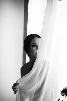 Black and white portrait of a woman peeking through a white curtain, creating a mysterious ambiance.