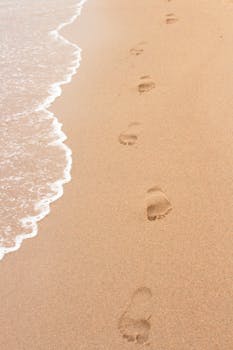 Footprints on a sandy beach beside ocean waves, perfect for summer themes.